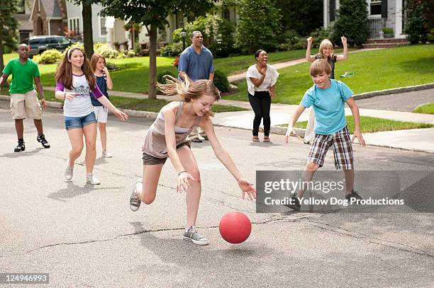 parents cheering kids in the background on the sidelines of the street, girl leaning over to grab kickball