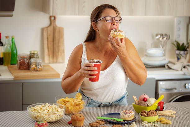 Plus size greedy woman enjoying eating a doughnut and drinking cola in her kitchen. Unhealthy eating concept