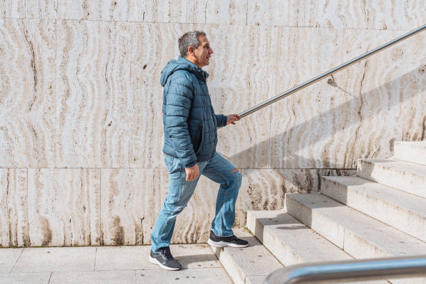 Portrait positive, cheerful middle-aged man, in warm blue jacket, jeans torn at knees, near marble wall, on steps with handrails