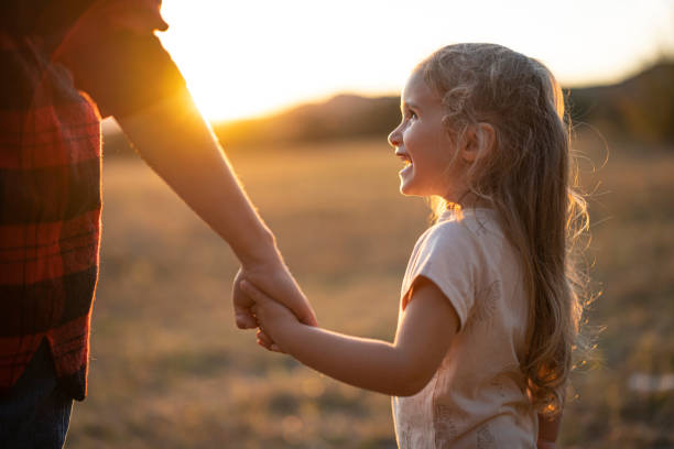A girl smiles radiantly at her mother while holding hands in a peaceful field, reflecting joy, innocence, and warmth in their mutual connection.