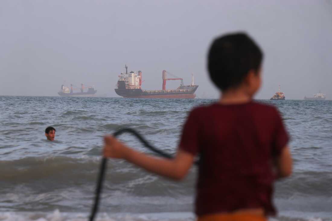 Children play in the water along the shore as a mix of bulk carriers, cargo ships, and service vessels sit offshore in the Strait of Hormuz off Bandar Abbas, Iran, Sunday, April 26, 2026.(Razieh Poudat/ISNA via AP)