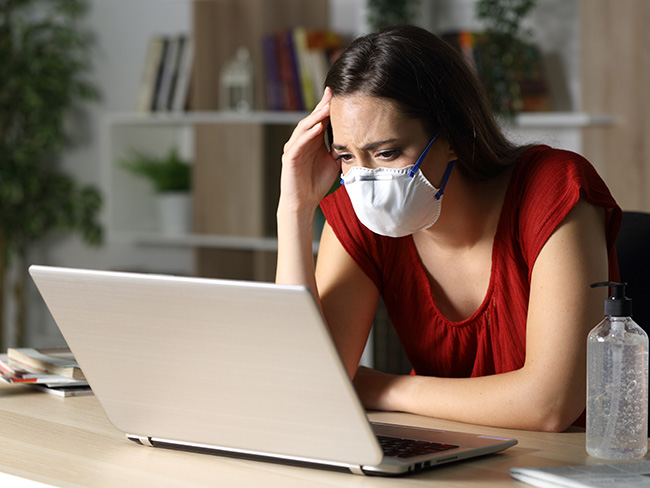 sad woman wearing mask looking at laptop and furrowing her brow
