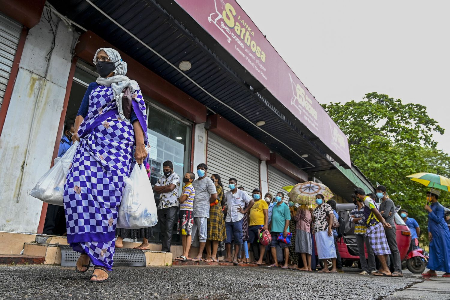 A woman carrying food bags walks pasts people standing in queue outside a state-run supermarket to buy essential food items in Colombo on September 3, 2021 as Sri Lanka began imposing price controls on essential food from September 3 after using a state of emergency to seize allegedly hoarded stocks of sugar and rice. (Photo by Ishara S. KODIKARA / AFP) (Photo by ISHARA S. KODIKARA/AFP via Getty Images)